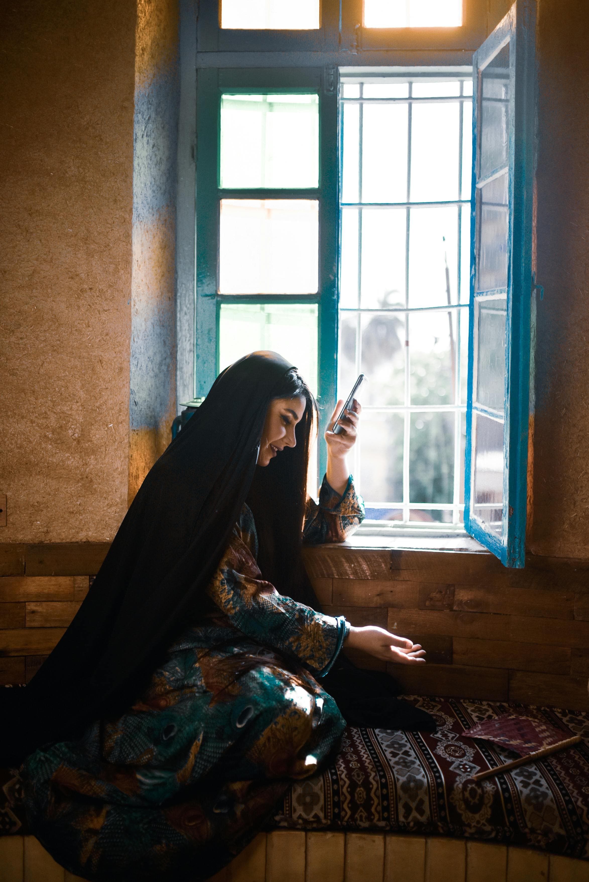 Afghanische Frau sitzt neben einem blauen Fenster und hält ihr Telefon